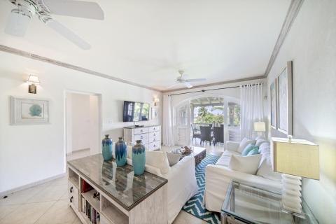 Open and airy living room in Sunshine View Penthouse at Sugar Hill Resort, Barbados, decorated with coastal-inspired decor, white furnishings, and a large television, flowing seamlessly onto the balcony with beautiful garden views.