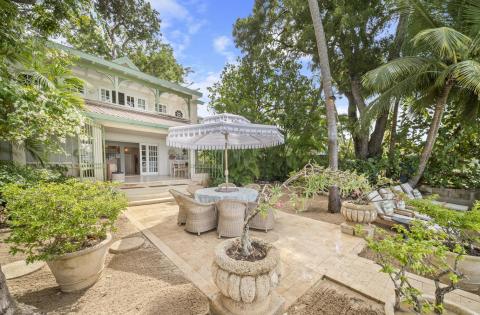 The outdoor patio of Seascape, a premier beachfront holiday villa in Barbados, features a shaded dining area with wicker furniture, surrounded by lush greenery and steps away from the white sandy beach.